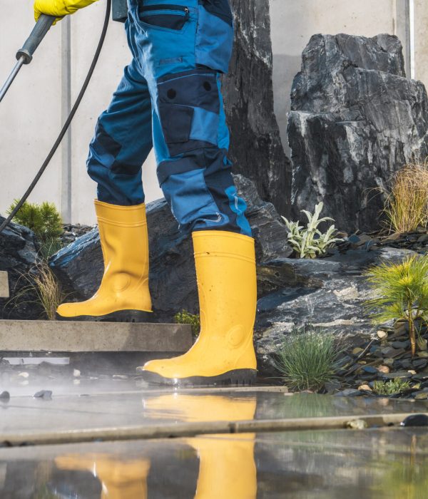 Men Wearing Yellow Rubber Boots Pressure Washing Architectural Concrete Elements in His Garden. Lower Body Close Up. Modern Concrete Backyard Stairs Cleaning.